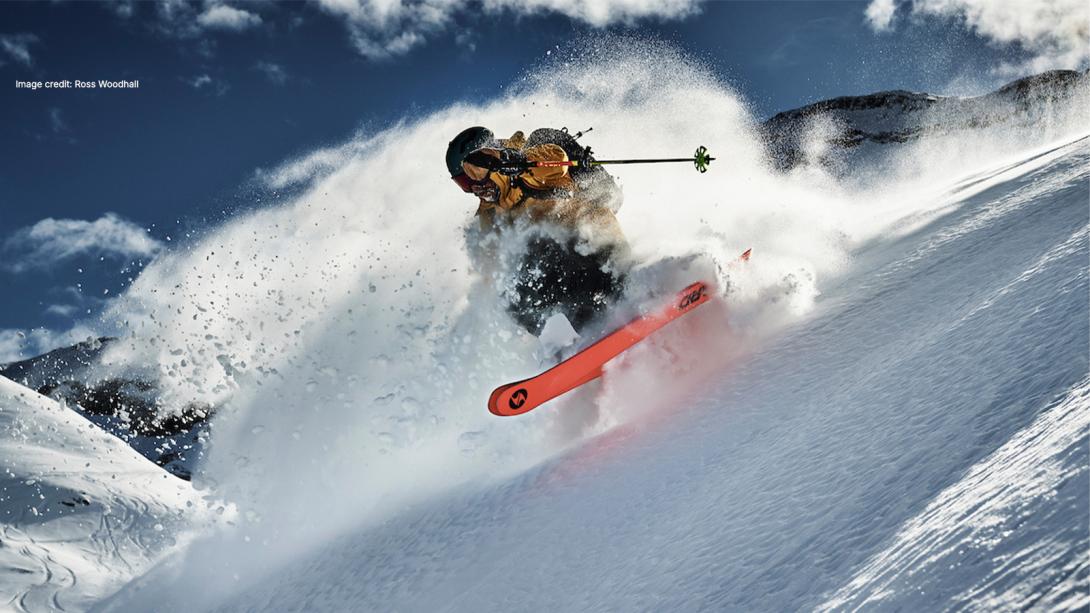 A skier in mid-action carves through fresh powder on a steep mountain slope, sending up a dramatic spray of snow under a bright blue sky. The skier wears a yellow jacket, black helmet, and goggles, with vibrant red skis visible through the snow. (Image credit: Ross Woodhall)