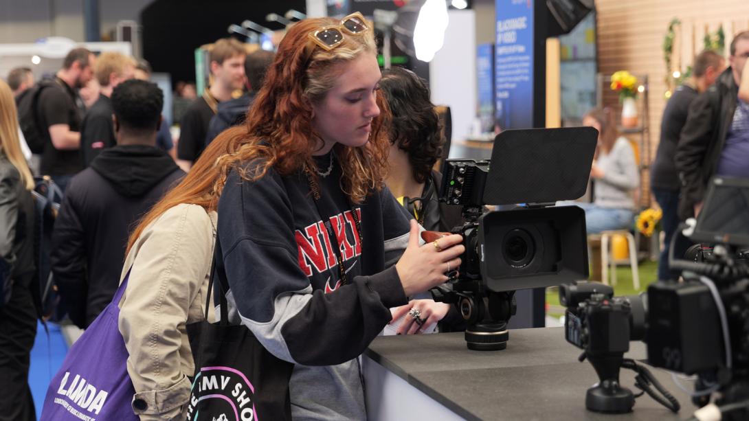 Young woman examining a professional video camera at a busy technology or media expo.    