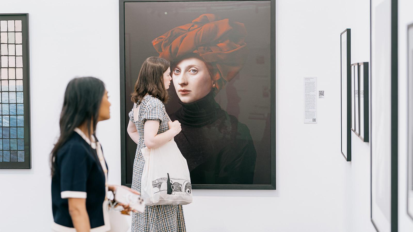 A visitor at a photography exhibition closely observes a large framed portrait of a woman in a red head wrap, displayed in a bright, white-walled gallery.