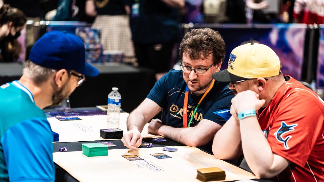 Three men sit at a table playing a trading card game, with one man in the centre pointing to a card on the table. 