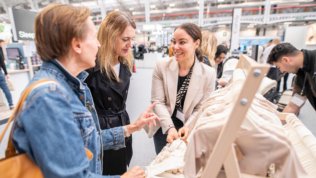 Two people discussing with an exhibitor the baby clothes they have on display 