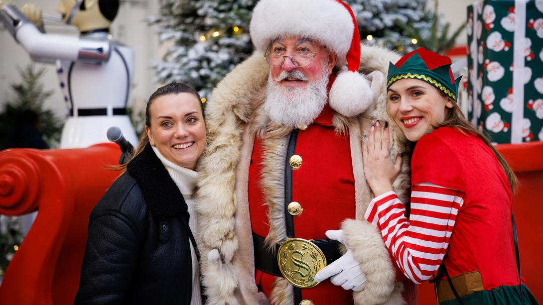 Santa Claus and an elf posing with a person next to a sleigh and festive decorations.