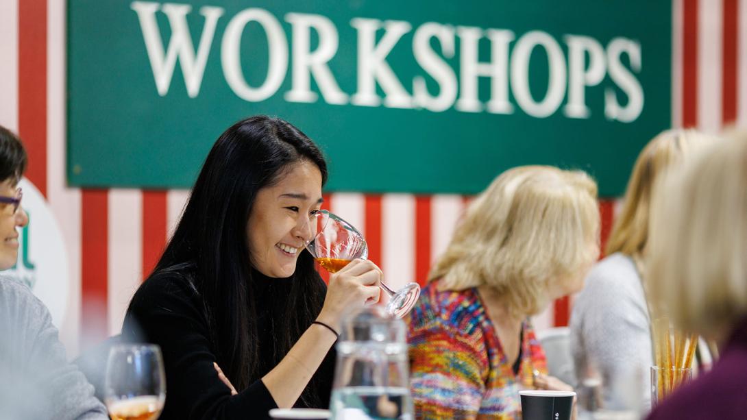 Person smiling while holding a wine glass at a table with others, under a 'WORKSHOPS' sign.