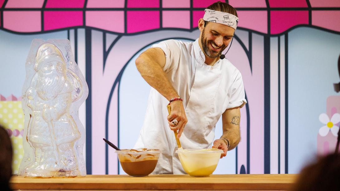 A person in a chef's outfit and headband smiles while stirring batter in a bowl at a baking competition, with a chocolate Santa Claus figure on the table.