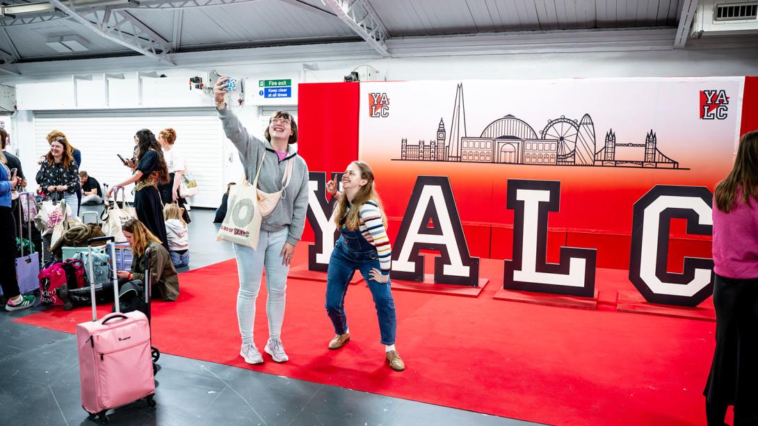 Two individuals taking a selfie in front of a large YALC (Young Adult Literature Convention) banner with city skyline graphics.