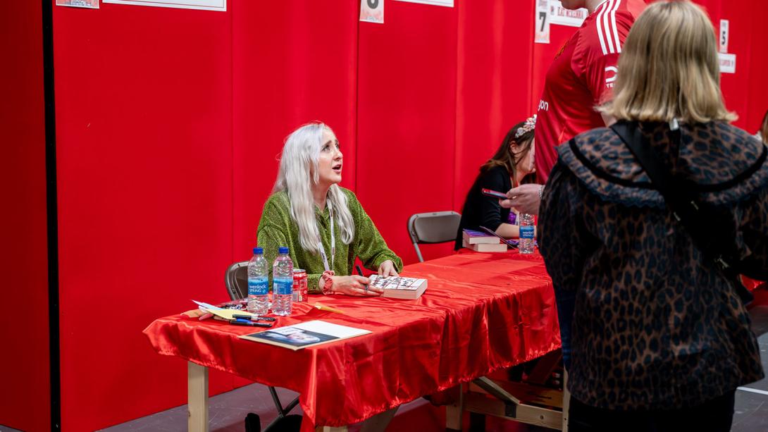 A person with long, white hair is seated at a signing table, interacting with attendees in front of a vibrant red background.