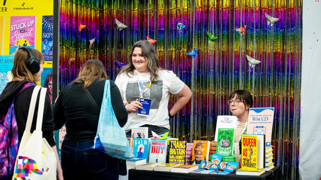 Two individuals stand behind a book stall at a literary festival, surrounded by colourful books and a vibrant, multicoloured tinsel background.