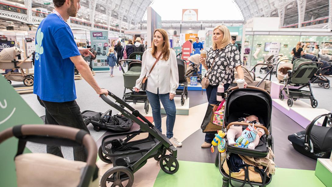 A sales representative demonstrates a stroller to two women at a baby products show, one of whom has a baby lying in a pushchair nearby.