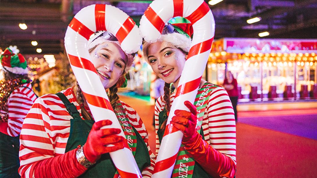 Two performers dressed as Christmas elves in red-and-white striped tops and green overalls hold oversized candy canes shaped into a heart, smiling at the camera.