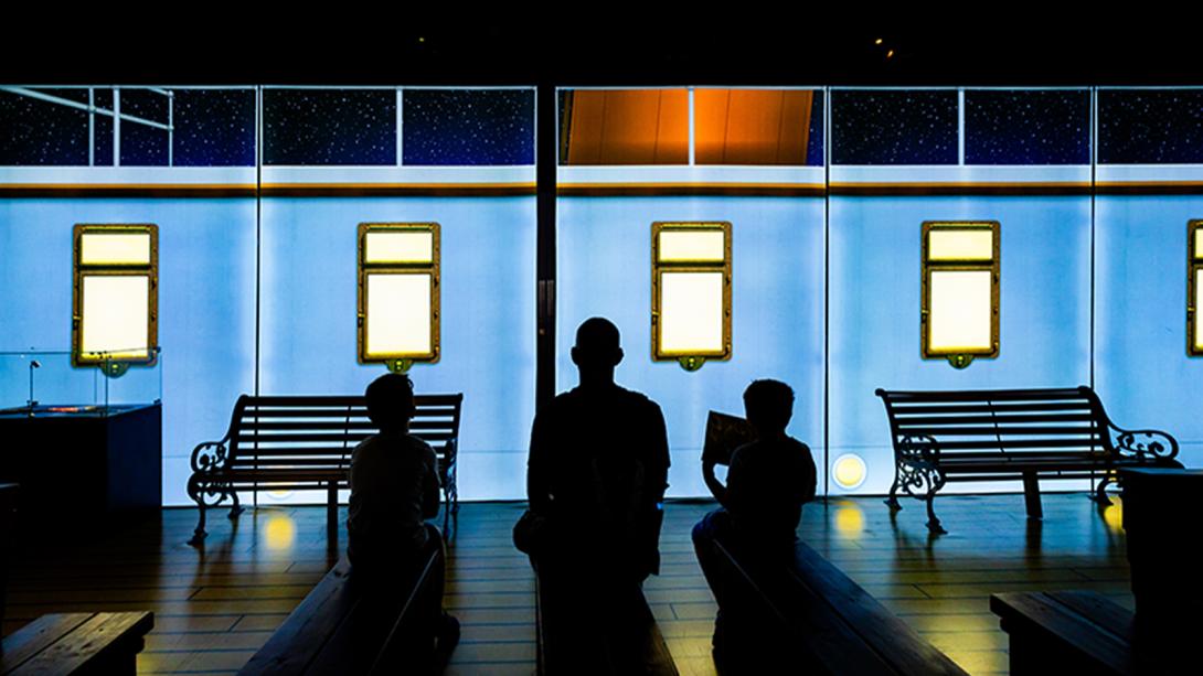 Silhouettes of three people sitting on benches in front of a brightly lit digital backdrop depicting a train with glowing windows and a starry night sky above.