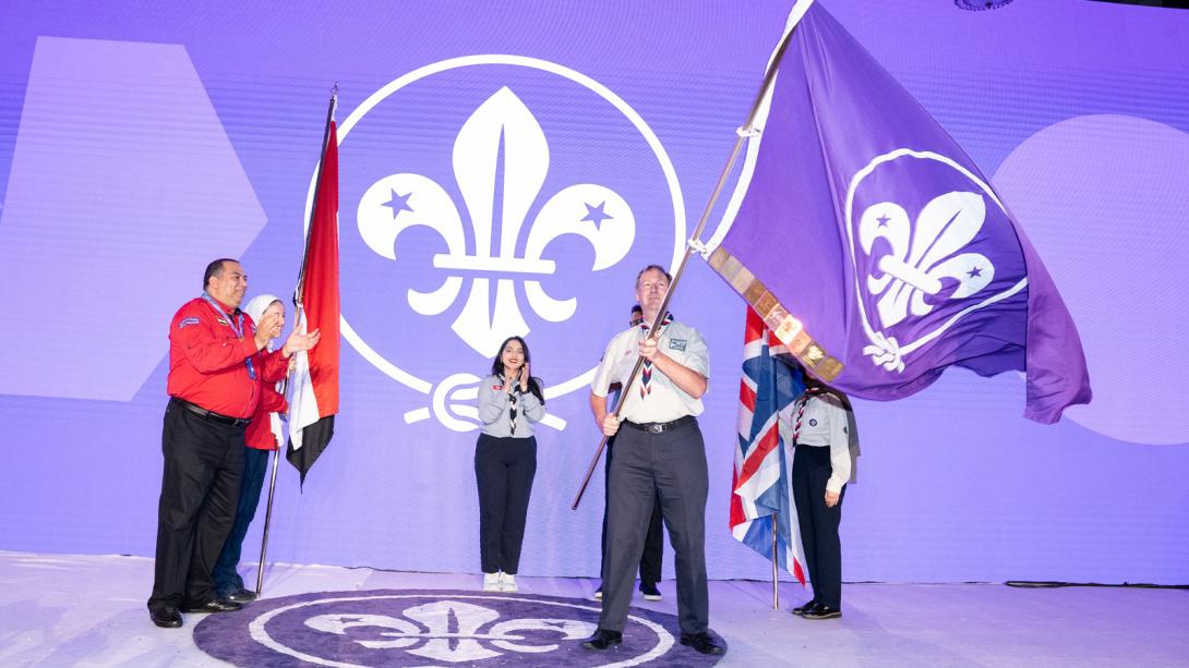 A group of Scouting representatives on stage holding flags, with a large purple backdrop displaying the Scouts fleur de lis symbol behind them.